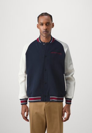 Chaqueta varsity en azul marino y blanca con cierre de botones, cuello y puños de canalé. Presenta mangas contrastantes y detalle del logo "TOMMY JEANS".