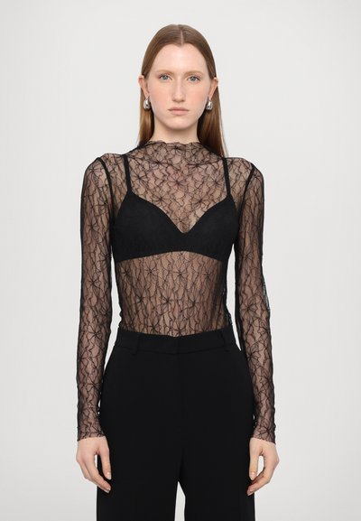 Schwarzes Spitzenoberteil mit langen Ärmeln, floralem Muster und hohem Kragen, das über einem schwarzen Bralette getragen wird, kombiniert mit schwarzen Hosen.