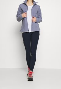 Grå fleecjacka med hög krage, med dragkedja fram och sidofickor, bärs över en vit skjorta och marinblå leggings.