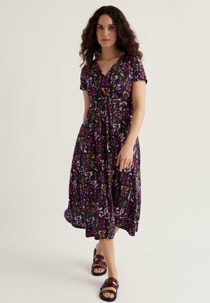 Kobieta o ciemnych, kręconych włosach, ubrana w granatową sukienkę midi w kwiaty z krótkimi rękawami oraz bordowe sandały, stojąca na tle jednolitego jasnego tła.