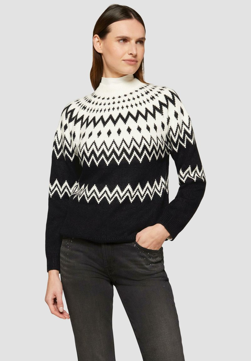 Gebreide trui met een hoge kraag, met een zwart-wit geometrisch patroon, zigzagontwerp en geribde manchetten. Draagt het met grijze jeans.