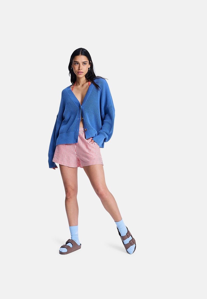 Femme portant un cardigan bleu surdimensionné, un short rayé rouge et blanc, des chaussettes bleues et des sandales marron, debout avec une main dans la poche.