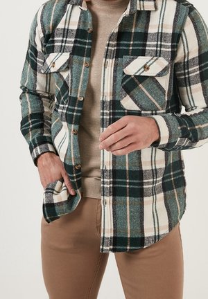 Mann trägt ein grün-weiß-beiges kariertes Hemd über einem beigen Pullover und brauner Hose, mit einer Hand in der Tasche und der anderen, die das Hemd richtet.