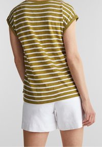 Persona vistiendo una camiseta de manga corta a rayas verdes y blancas y pantalones cortos blancos, mostrada de espaldas contra un fondo liso.