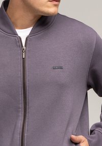 Fioletowa bluza z zamkiem na całej długości, z wysokim kołnierzem oraz małym logo marki na klatce piersiowej. Gładka tekstura materiału i luźny krój.
