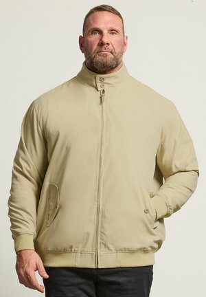 Mand med skæg iført beige lynlåstrøje med knappet krave, den ene hånd i lommen, stående mod en ensfarvet off-white baggrund.