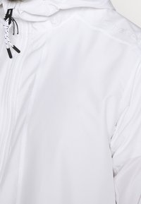 Chaqueta blanca ligera e impermeable con una textura suave, cierre de cremallera, capucha con cordón y detalles en negro contrastantes.