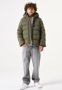 Chaqueta acolchada verde oliva con cremallera, capucha y bolsillo en el pecho; combinada con jeans grises y zapatillas blancas. Tejido texturizado, diseño aislante.