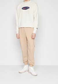 Ljusbeige sweatshirt med en marinblå logotyp, matchad med ljusbeiga joggers med sidodekaler, samt vita sneakers med grå sulor.