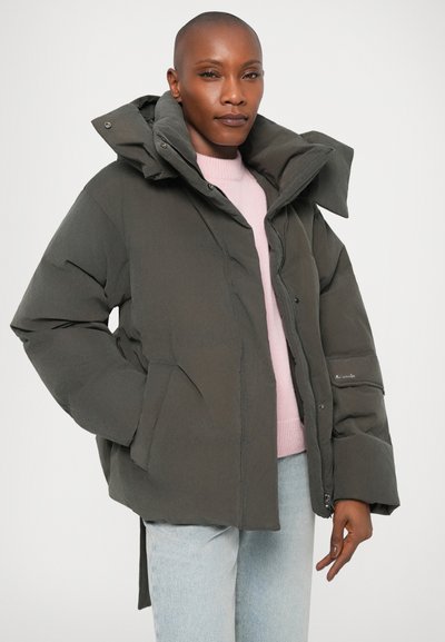 Persoon in een grijze oversized pufferjack over een lichtroze trui en lichtblauwe jeans, tegen een effen achtergrond.