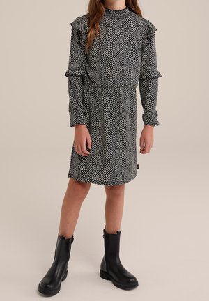 Fille portant une robe à motifs géométriques noir et blanc avec manches longues et des bottines noires sur un fond uni.