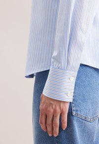 Camicia a righe blu chiaro con righe bianche e blu scuro. Presenta un polsino abbottonato e un orlo dritto, abbinata a pantaloni in denim blu.
