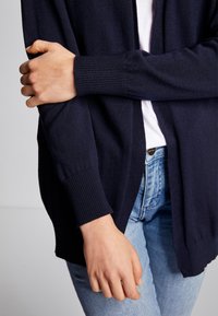 Mörkblå stickad cardigan med avslappnad passform, ribbade mudd, och öppen framsida, bärs över en vit skjorta och ljusblå jeans.