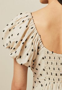 Beige klänning med puffärmar, svarta broderade prickar och en smockad bakpanel. Mjuk tyg med en strukturerad finish, med en djup halsringning.