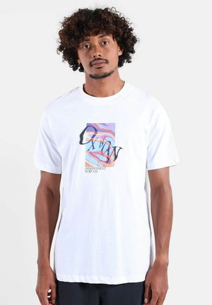 Homme aux cheveux bouclés portant un t-shirt blanc avec un motif de surf abstrait coloré et le texte « Independent Surf Co. », debout devant un fond uni.
