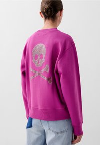 Pulover fucsia cu un grafic mare de craniu și oase încrucișate din sclipici pe spate. Manșete și tiv cu dungi, material moale din bumbac.