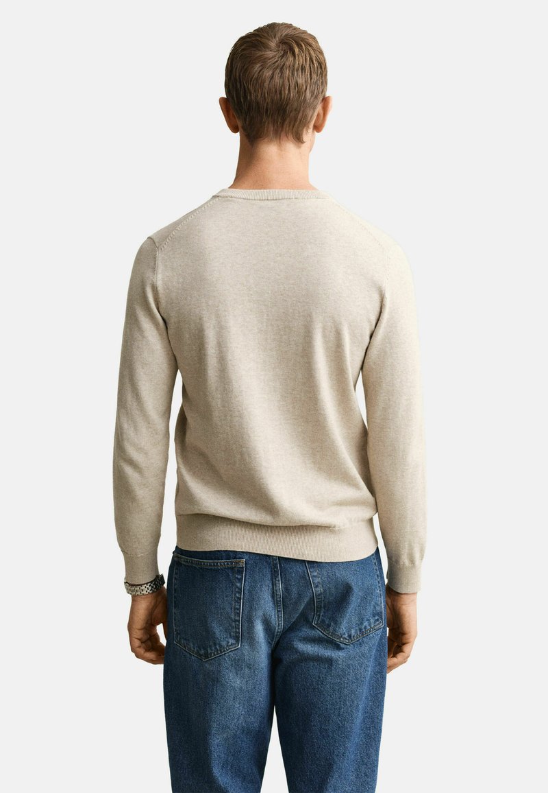 Mann trägt einen hellbeigen Langarmpullover und blaue Jeans, steht mit dem Rücken zur Kamera vor einem schlichten weißen Hintergrund.