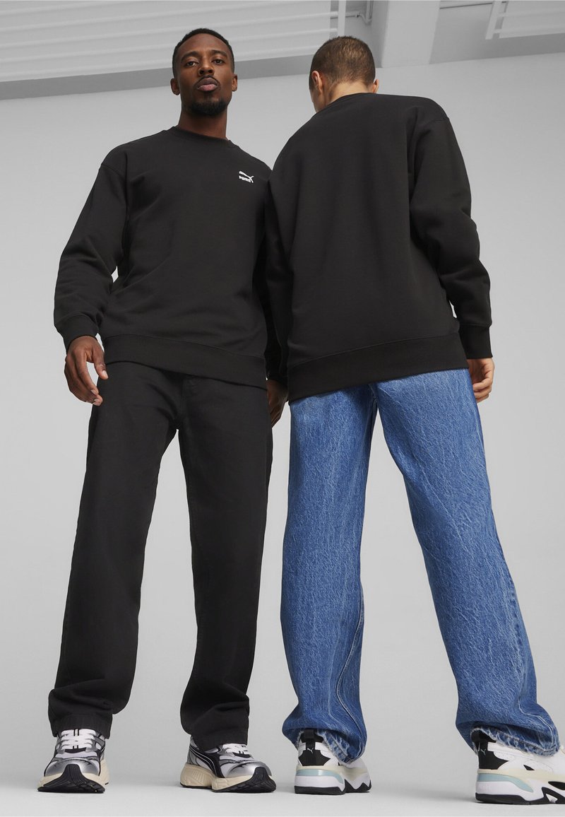 Felpa nera con collo a giro, abbinata a pantaloni neri e jeans larghi blu. Entrambi indossano sneakers multicolori. Visibile un minimo di branding.