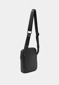 Musta olkalaukku, joka on valmistettu sileästä materiaalista, neliömäinen muoto, jossa on säädettävä hihna, jossa on kohokuvioitu brändäys ja vetoketjukiinnitys.