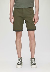 Oliwkowe szorty wykonane z mocnej tkaniny, z podwiniętymi brzegami i kieszeniami z przodu, w połączeniu z zielonymi trampkami za kostkę z białymi sznurowadłami.