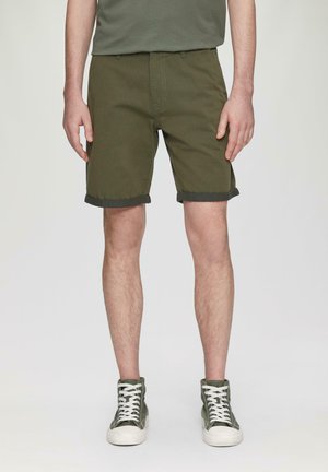 Olivgrüne Shorts aus strapazierfähigem Stoff, mit aufgeschlagenen Säumen und Fronttaschen, kombiniert mit grünen High-Top-Sneakern mit weißen Schnürsenkeln.