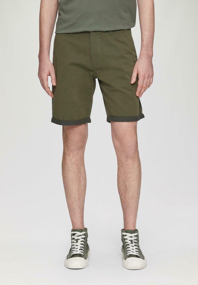 Oliwkowe szorty wykonane z mocnej tkaniny, z podwiniętymi brzegami i kieszeniami z przodu, w połączeniu z zielonymi trampkami za kostkę z białymi sznurowadłami.