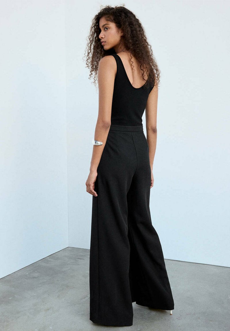 Femme aux cheveux bouclés portant un haut noir sans manches et un pantalon noir à jambes larges, debout contre un mur blanc avec un bracelet en argent.