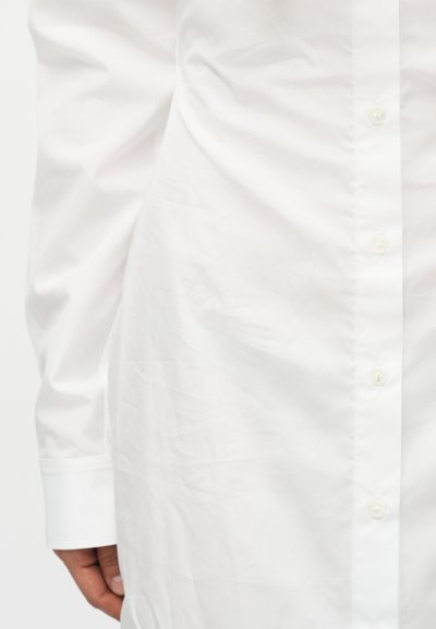 Κοντινό στο σώμα ενός ατόμου που φοράει λευκό πουκάμισο με μακριά μανίκια, ορατά κουμπιά και μανσέτα, το χέρι ακουμπά στο πλάι.