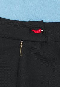 Pantalones negros con una cremallera con un tirador dorado y un pequeño acento de pimiento chili rojo en la cinturilla. Textura de tela suave.