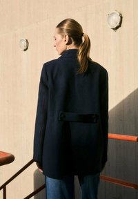 Marineblauwe wollen blazer met een gestructureerde silhouette, enkele knoop aan de achterkant, en een gelapte kraag, gecombineerd met lichtblauwe spijkerbroek.