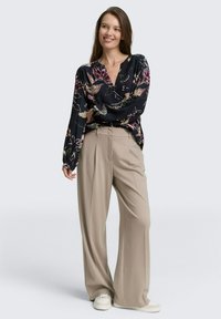 Kvinna som ler, iklädd en långärmad blus med svart blommönster, beige vida byxor och vita sneakers, stående mot en neutral bakgrund.