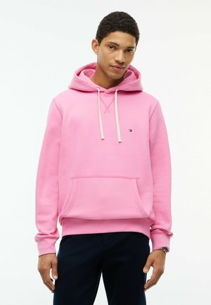 Junger Mann trägt einen pinken Hoodie mit weißen Kordeln und dunklen Hosen, steht vor einem schlichten weißen Hintergrund.
