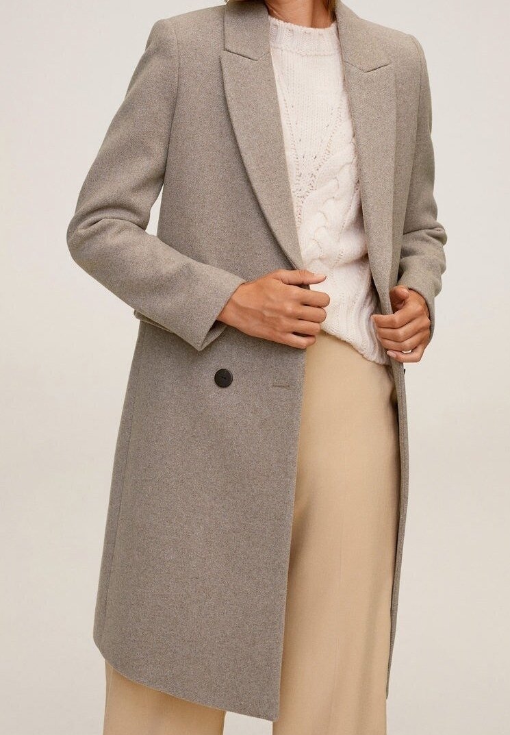 Personne portant un manteau en laine marron clair par-dessus un pull blanc à torsades et un pantalon beige, tenant les revers du manteau avec les deux mains.