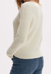 Pull en tricot couleur crème avec un motif texturé, doté d'un col bateau et de manches longues, associé à un jean bleu foncé.