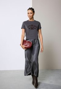 Graues, kurzes T-Shirt mit kastanienbraunen Text, kombiniert mit einem langen grauen Satinrock. Hält eine burgunderfarbene Clutch; trägt schwarze High-Heels.