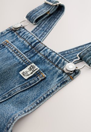 Džinsinės petnešos šviesiai mėlynos spalvos, su reguliuojamomis juostelėmis, metalinėmis sagtimis ir etiketė, kurioje ant priekinio kišenės vaizduojami žiedai.