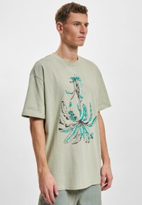 Světle zelené oversized tričko z bavlny s modro-černým květinovým grafickým designem. Má kulatý výstřih a krátké rukávy.