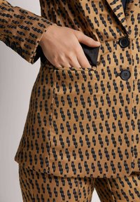 Tan blazer met zwart abstract patroon, voorzien van een notch revers, drie knopen en een zijzak, geaccentueerd tegen een gladde stof.