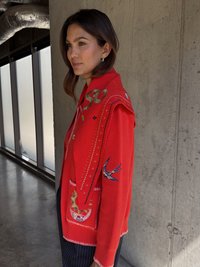 Piros gyapjú kardigán bonyolult hímzésekkel, úszómadár mintákkal és színes motívumokkal, dekoratív varrással és zsebekkel.