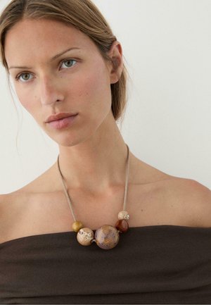 Donna con capelli castano chiaro indossa un top scuro scollato sulle spalle e una collana con cinque grandi perle rotonde in toni naturali su una sottile catena.