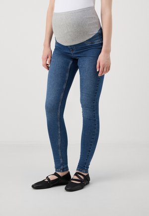 Graviditets skinny jeans i mørkeblå denim med en grå elastisk livlinje. Funksjoner inkluderer en tettsittende fasong og ankellengde. Sorte flate sko.