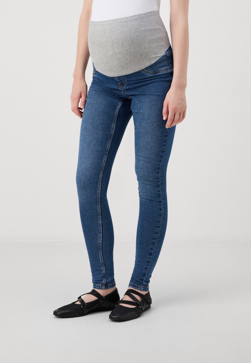 Sötétkék denim anyagú terhes skinny farmer, szürke gumírozott derékkal. Jellemzői között szerepel a testhezálló forma és a bokáig érő kivitel. Fekete lapos cipők.