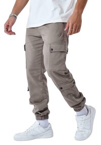 Pantalones cargo beige con puños ajustados, múltiples bolsillos laterales y botones a presión negros, acompañados de zapatillas blancas y calcetines hasta la mitad de la pantorrilla.