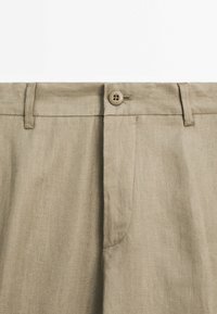Pantalon en lin beige en gros plan montrant la ceinture avec fermeture par bouton, boutonnière et passants de ceinture.