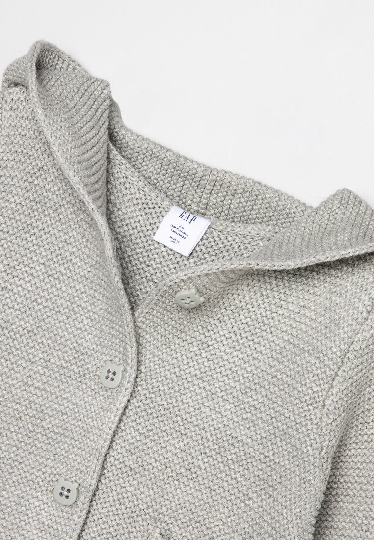 Cárdigan de punto gris claro con cuello de chal, que presenta botones grandes y un patrón texturizado. Etiqueta de cuidado visible en el interior.