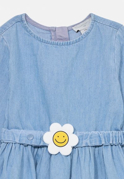 Ανοιχτό μπλε τζιν φόρεμα με στρογγυλή λαιμόκοψη, μαζεμένη μέση και λευκή διακόσμηση σε σχήμα λουλουδιού που έχει ένα κίτρινο χαμογελαστό πρόσωπο.