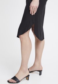 Vestito nero con un alto spacco laterale, in tessuto leggero e dal design minimalista e elegante. Abbinato a sandali neri con tacco e punta aperta.