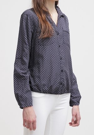 Kvinde iført en marineblå mønstret skjorte med lange ærmer og elastisk kant, kombineret med hvide bukser, stående mod en ensfarvet baggrund.