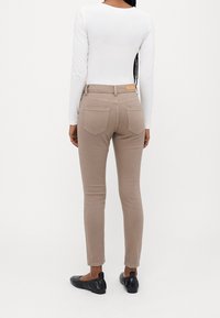 Jeans skinny beige con cintura de tiro medio, que cuentan con dos bolsillos traseros y una etiqueta de cuero marrón en la parte trasera. Llevados con una blusa blanca de manga larga.