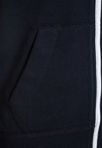 Donker marineblauwe sweatshirtstof met een voorzak, gestructureerd oppervlak, zichtbare stiksels en een witte ritsdetails langs het midden.